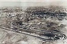 Image en noir et blanc. Au fond, la ville de Los Angeles. À l'avant-plan, un immense ovale bordé de petites maisons de plain-pied, qui forment le village olympique ; l'intérieur de l'ovale contient trois bâtiments en longueur et de la verdure.