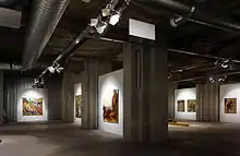 Photographie en couleurs de la salle principale vue d'un angle, des piliers de béton qui la parcourent et de quelques œuvres.