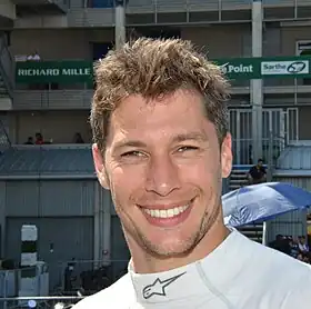 Photographie d'un homme brun, vu en gros plan, habillé dans un polo blanc, de trois-quarts.