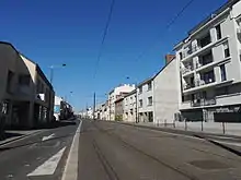 Rue bordée d'immeubles de trois ou quatre étages, des rails de tramway parcourent le centre de la voie