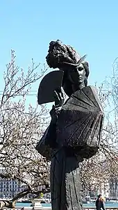 Photo en couleurs d'un buste sur un pilier représentant une femme sculptée portant un chapeau à plumes couvrant ses cheveux nattés.