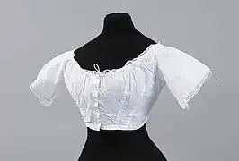 Cache corset pour crinoline en cotonnade blanche. Décolleté forme bateau, petits mancherons soulignés de dentelle à l’aiguille. Boutonnage milieu devant par 5 boutons recouverts et une agrafe.   Vers 1860.