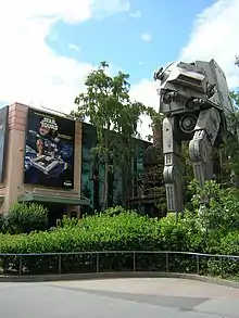 Forêt avec des huttes dans les arbres, un grand véhicule à quatre pattes, et un bâtiment avec un poster.