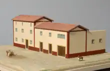 Photographie de la maquette d'une maison gallo-romaine. Les murs sont beige et rouge, le toit imite des tuiles. Il y a deux étages au bâtiment principal, et les portes sont en bois.