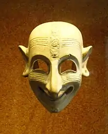 Photographie d'un masque strié horizontalement, posé sur un fond doré, diadème sur le front, nez prédominant, rire sardonique.