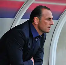 Homme brun, assis sur un banc, vu de profil, regardant fixement devant lui.