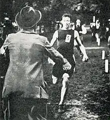 Un homme portant le dossard 3, au premier plan un homme en costume avec un chapeau lui ouvrant les bras.