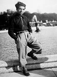 Photographie en noir et blanc d'un homme posant devant une fontaine, un pied sur le rebord.