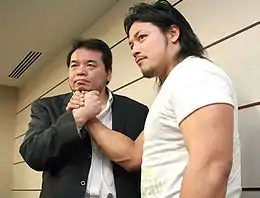 Mitsuharu Misawa en costume serre la main de Gō Shiozaki qui porte un T-Shirt blanc.