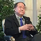 Photographie en plan taille d'un quinquagénaire en costume-cravate, assis avec les mains jointes devant lui.