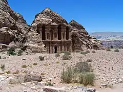 Vue de loin d'un monument sculpté dans la roche. Une esplanade de roche et de sable se trouve en avant plan dans lequel deux promeneurs admirent le paysage.