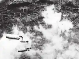 Photographie en noir et blanc prise en contre-plongée depuis un bombardier en train de déverser des bombes sur une ville.