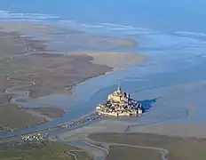 Vue aérienne du mont Saint-Michel : le rocher (sans les bâtiments) présente une base grossièrement elliptique de 950&nbsp;m de circonférence pour une superficie de 28&nbsp;ha, et mesure 91&nbsp;m de haut.