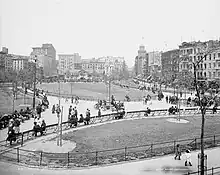 Photographie en noir et blanc de personnes marchant dans les allées d'un parc public, avec en arrière-plan des immeubles et bâtiments hauts.
