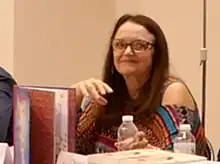 Photo en couleurs d'une femme à longs cheveux châtain foncé, avec lunettes, derrière des livres.