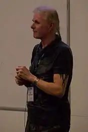 A neutrally looking elderly man with gray hair and a horse tail. He has a tattoo on the left upper arm, as well as a bracelet and a ring.