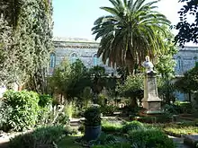 Buste au sommet d'une colonne, dans un jardin exotique. En arrière-plan, un bâtiment d'un étage.