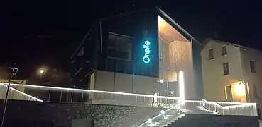 Photographie de la face sud de l'office de tourisme (de nuit), avec un éclairage bleu et des maisons dans le parage.