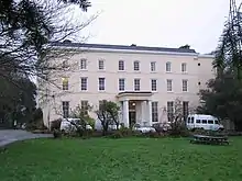 Photo d'un bâtiment rectangulaire de trois étages à la façade saumon, avec de nombreuses fenêtres. L'entrée est sous un porche soutenu par quatre colonnes blanches