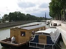  Photo couleurs. Deux embarcations sont arrimées aux abords de l’écluse d’Orléans. Un est en bois, avec une grosse cabane, l’autre est plus grande. En arrière, on perçoit les escaliers de pierre qui permettent aux plaisanciers de regagner le quai du fort Alleaume et les bords de Loire d’Orléans. Plus loin, on voit les mats de la capitainerie d'Orléans.