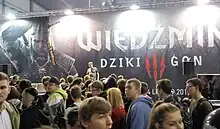 Photographie couleur d'une foule de passants devant un stand aux couleurs du jeu.