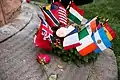 Photo agrandie de la base grise du monument. Une couronne de houx verte et entourée de plusieurs drapeaux de divers pays et de fleurs.