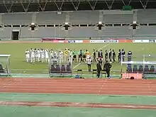 Dans un stade de football, deux équipes se tiennent alignées et applaudissent.