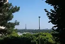 Photographie en couleur de végétation et de quelques bâtiments au premier plan et d'une tour à l'arrière-plan.