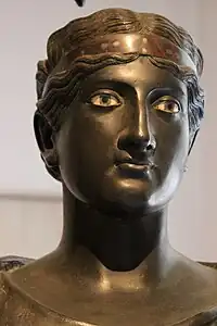 L'une des 5 péplophores (vêtues d'un péplos). Plusieurs semblent s'habiller. Celle-ci porte un bandeau dans les cheveux, composé de plusieurs métaux, choisis pour leurs couleurs.