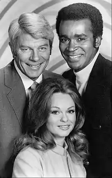 Photo en nir et blanc de deux hommes et une femme, tous trois souriants.