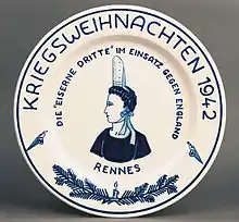 Plat en faïence blanche. Au centre du plat, un buste de Bigouden représenté au trait bleu, de trois quart, tête de profil gauche. Sur le marli, de couleur bleue, deux branches de sapin et une bougie allumée sont encadrés par deux oiseaux de proie stylisés portant un écusson.