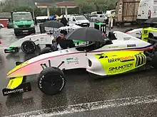 Photographie d'une monoplace de F4 blanche et jaune fluo, vue de profil gauche, devant une autre voiture, à l'arrêt, sur une piste humide.