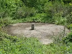Une dalle en béton circulaire surmonté d'une borne est entouré par la végétation composé de broussailles et d'arbustes.