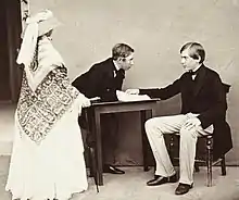 la reine Victoria debout et de dos, vêtue d'une toilette claire, recouverte par un châle brodé et coiffée par un chapeau de paille entouré d'une étoffe nouée regarde ses deux cousins, Louis semi-incliné sur une table regardant son frère Pierre assis et tenant quelques feuilles de papier, les deux princes portent une redingote de couleur foncée