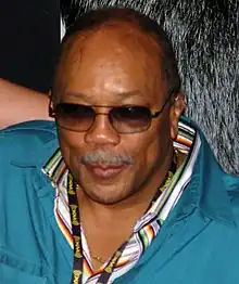 A man in a blue jacket looks ahead. The balding male's dark sunglasses conceal his eyes.