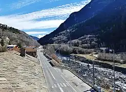 Photographie d'une route franchissant une agglomération avec des maisons aux toits de pierres et une rivière agitée à droite ; on aperçoit un rond-point tout au fond ainsi que des passages piétons et parkings.