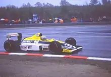 Photo d'une monoplace jaune, blanche et bleue, vue de côté droit, sur une piste détrempée.