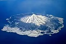 Photo couleur d'un volcan aux pentes enneigées, sur une île au milieu d'une mer bleue, sous un ciel bleu.