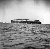Photo en noir et blanc prise depuis un bateau avec la mer et au centre le rocher