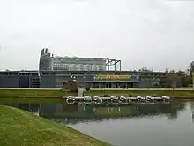 Photographie du Jardin du Monde, montrant un bâtiment bas incurvé, à structure de métal et panneaux pleins de couleur gris foncé, avec en son centre une serre enserrée dans un réseau métallique. Devant s’étale, entouré de pelouses, un plan d’eau avec un ponton et des petits bateaux à quai.