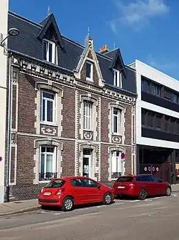 Cliché numérique couleur. Devant une maison de la fin du XIXe&nbsp;siècle et un immeuble du XXe&nbsp;siècle de trois étages stationnent deux voitures rouges.