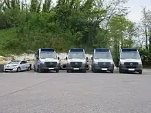 Photographie en couleurs de cinq véhicules garés côte-à-côte, vus de face : une voiture électrique blanche et quatre minibus bleu, blanc et noir. Il sont stationnés devant un mur en pierre blanches et des arbres.