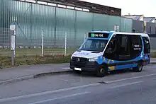 Photographie en couleurs d’un minibus bleu, blanc et noir, stationné devant des bâtiments à l'aspect moderne