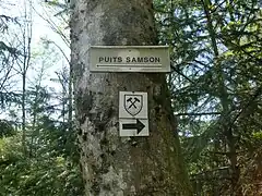 Panneau portant la mention « puits Samson » et une flèche dirigée vers la gauche tandis que le sentier minier est indiqué vers la droite.