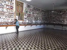 Photographie en couleurs. Intérieur d'une salle, dallée avec des triangles noir et blanc ; sur les murs, de nombreux ex-voto sont accrochés, que consulte une femme.