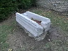 Sarcophage pris en contre-plongée, en pierre blanche, posé sur la terre. La paroi en largeur est endommagée, celle en longueur est rainurée.