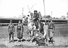 Photographie en noir et blanc représentant un cavalier entouré de quatre personnes, dont une assise par terre.