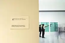Exposition d'un requin dans une eau verte, un mur devant avec une affiche indiquant que l’œuvre, Sensation des Young British Artists, a d'abord été exposée à l'Académie royale des arts de Londres