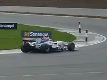 Photographie d'une monoplace blanche, bleue et rouge, vue de trois-quarts dos droit, à l'amorce d'un virage vers la gauche.