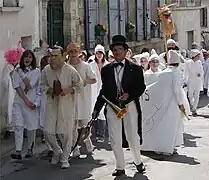 Un homme à lunettes, habillé d'un haut-de-forme et d'une queue-de-pie, dirige un cortège de Soufflaculs, tous vêtus de chemises de nuit blanches et de bonnets de coton blanc, avec à leurs mains des soufflets.